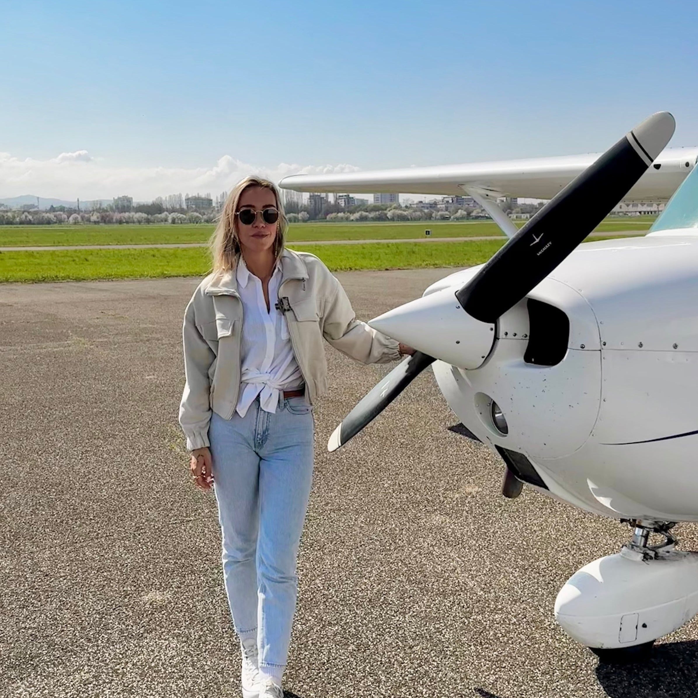A woman wearing a light beige jacket, white shirt, and light blue jeans walks beside a white propeller airplane on an airport tarmac under a clear blue sky.