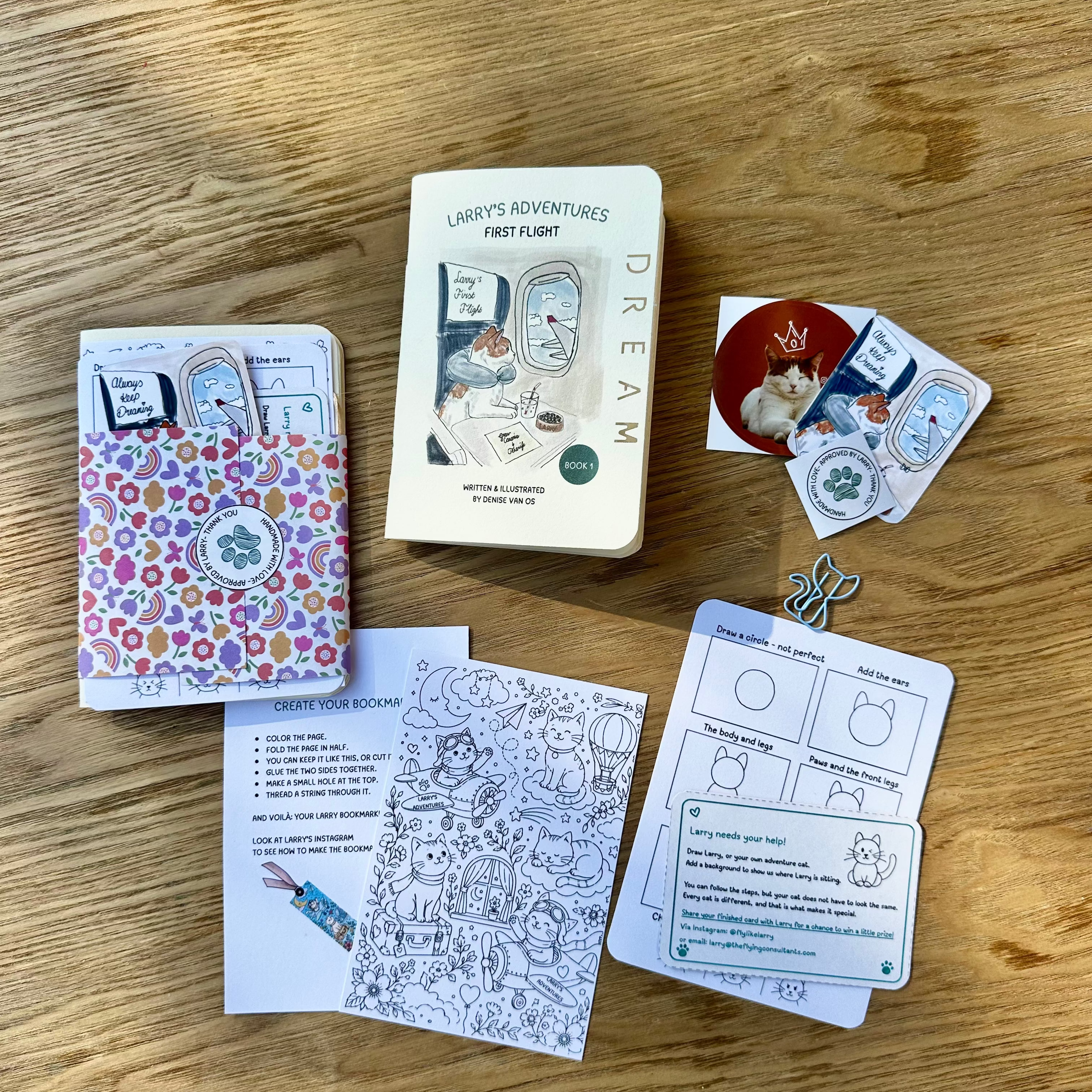 A children's activity book  by 'Larry's Adventures' arranged on a wooden tabletop. The set includes a small notebook with a colorful floral pattern, a white hardcover book, several printed cards, and a small sticker or charm.