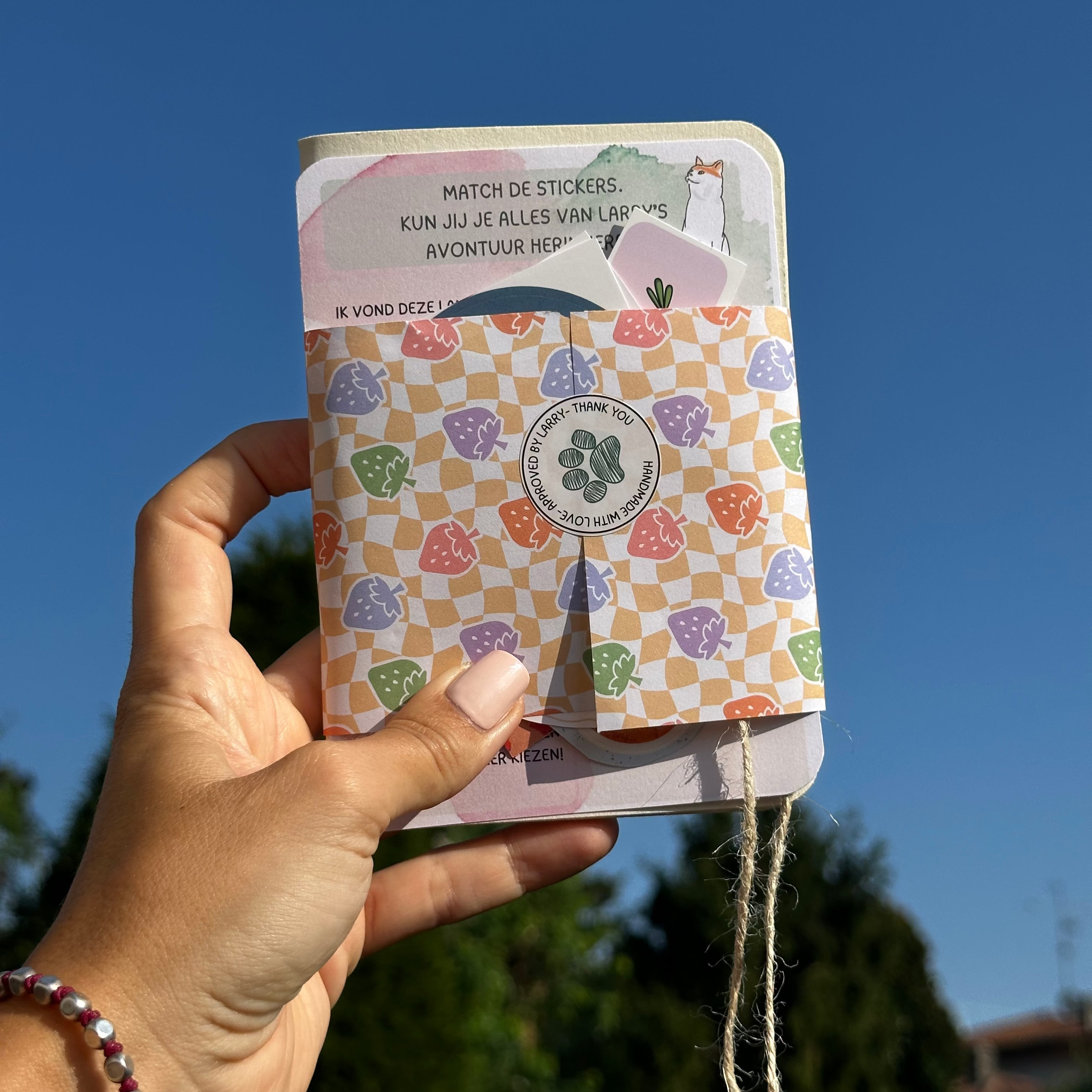 A hand holding a small notebook or journal outdoors against a blue sky. The cover has a pastel multicolor checkered pattern with purple, pink, green, and orange shapes on a light background, and a small flap reveals a sticker sheet with Dutch text.